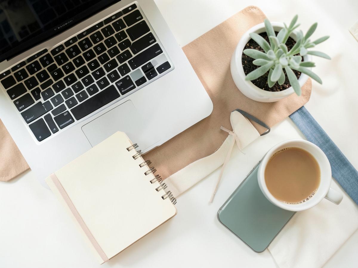 A bright desk with a laptop, notebook, phone, coffee, and a sage-green plant
