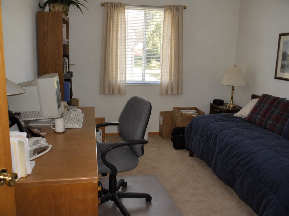 A small spare bedroom set up as a simple home office with an old computer, a desk, file folders, and a bed